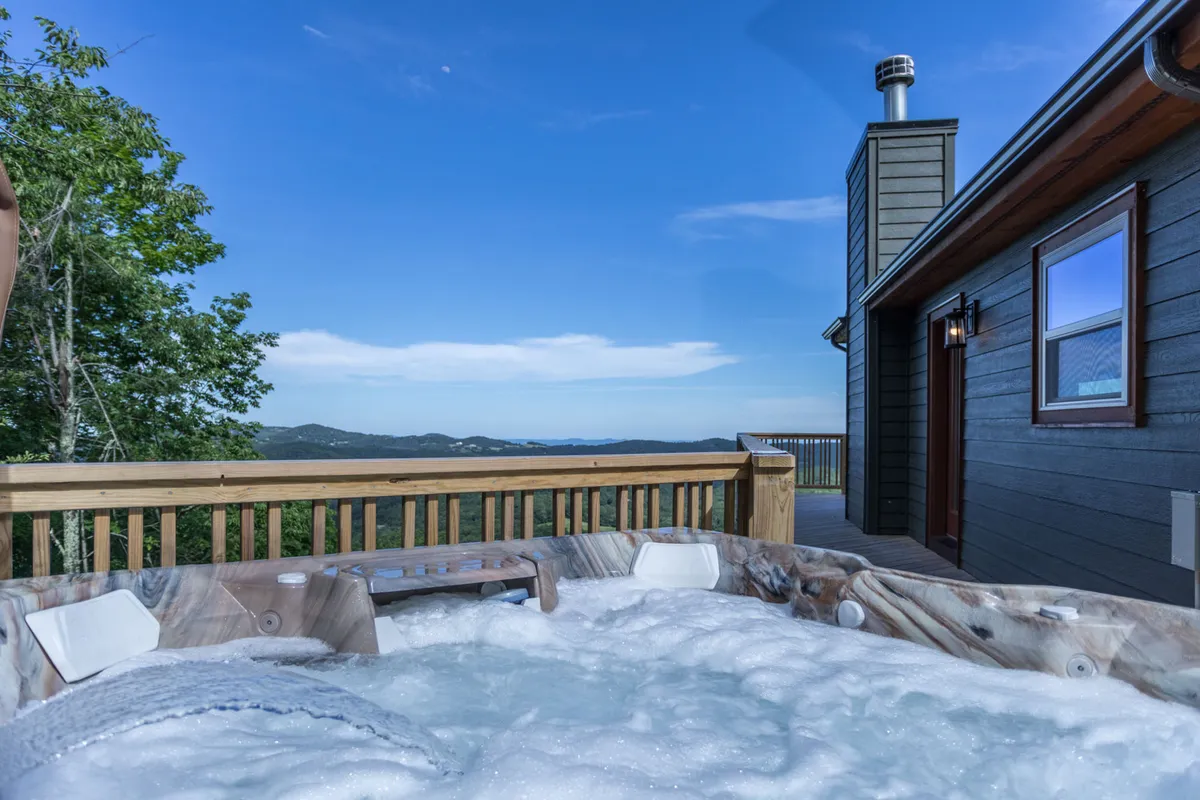 A serene mountain-facing hot tub sits on a wooden deck, surrounded by sturdy railings. The panoramic view stretches to distant Blue Ridge peaks, and the placement ensures privacy while inviting guests to relax and take in the vista. The hot tub's steam suggests year-round enjoyment, and a portion of the wrap-around deck is visible, emphasizing the connection to the outdoors.
