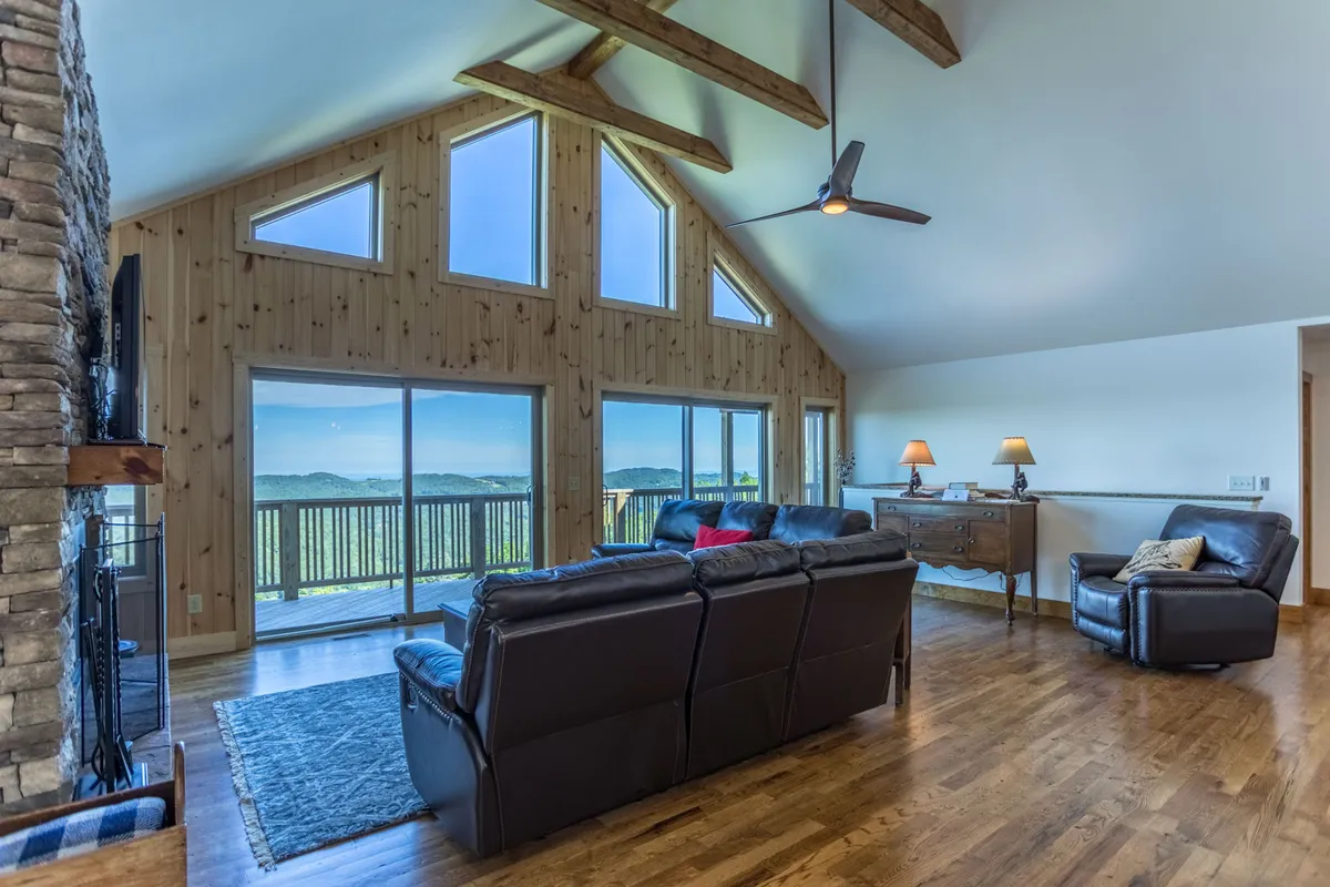 A spacious Great Room featuring a stone fireplace, plush leather seating arranged to face large windows, and exposed wooden beams on the ceiling. The expansive windows frame breathtaking mountain views, and the warm lighting creates an inviting atmosphere ideal for relaxation and gatherings.