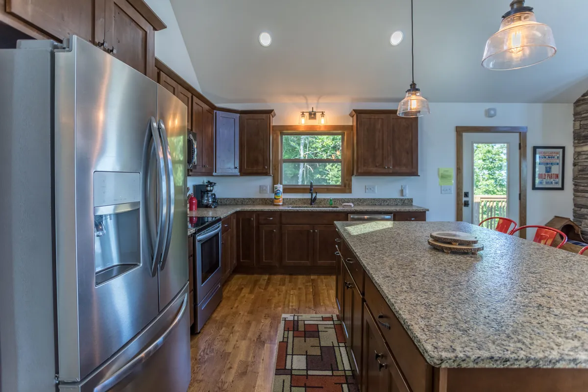 A modern kitchen with stainless steel appliances, granite countertops, wooden cabinetry, and an island with seating beneath a large window. The space feels bright and welcoming, suitable for casual gatherings or preparing gourmet meals.