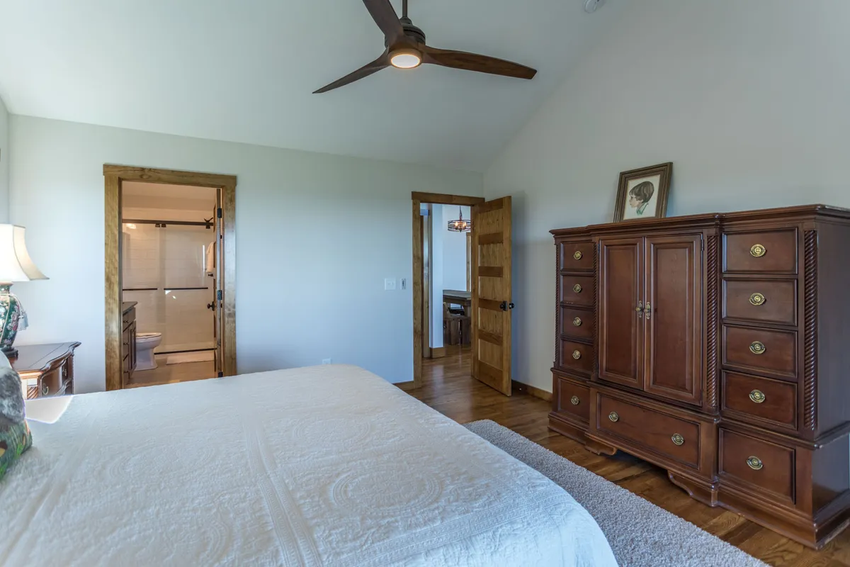 A comfortable bedroom with a neatly made bed featuring a quilted cover, a wooden dresser for ample storage, and an open door leading to a glimpse of the ensuite bathroom. Soft, natural lighting enhances the serene and welcoming mood of the space.